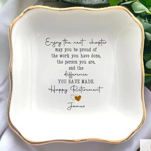 May include: A white ceramic dish with a gold trim and a square shape. The dish has a retirement message in black script: "Enjoy the next chapter...Happy Retirement James."