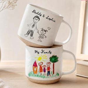 May include: Two white ceramic mugs stacked on top of each other. The top mug has the text "Daddy & Sophia" and a drawing of a father and child. The bottom mug has the text "My Family" and a drawing of a family with a house and tree.