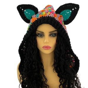 Op de afbeelding: Een zwarte gehaakte capuchon met grote kattenoren. De oren zijn zwart met teal en de capuchon is veelkleurig. De capuchon wordt gedragen over lang, zwart krullend haar. Handgemaakt.