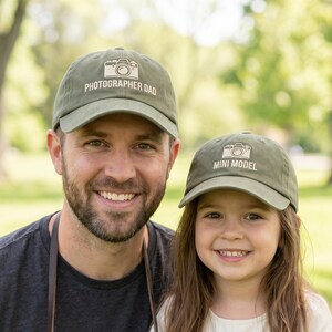 May include: Two olive green baseball caps with a camera graphic. One cap reads "Photographer Dad," the other reads "Mini Model." A man and a child are wearing the caps, smiling at the camera. The man is holding a vintage camera.