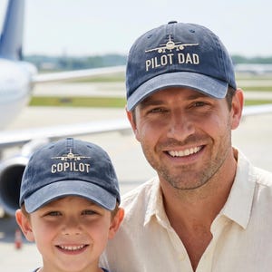 May include: Two navy blue baseball caps with white text and a plane graphic. One cap says "PILOT DAD" and the other says "COPILOT". The caps are worn by a man and a child, with an airplane in the background.