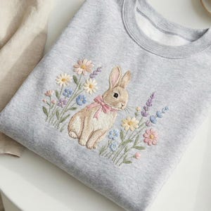 May include: A light gray sweatshirt featuring an embroidered design of a bunny with a pink bow, surrounded by colorful flowers. The design includes daisies, lavender, and other wildflowers. The sweatshirt is folded on a white surface.