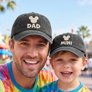 May include: Two black baseball caps with the text "DAD" and "MINI" in white, each featuring a Mickey Mouse silhouette. The caps are worn by a man and a child, both smiling. They are wearing tie-dye shirts.