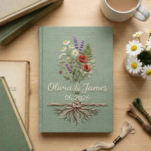 May include: A sage green journal with an embroidered floral design featuring daisies, poppies, and lavender. The cover is inscribed with "Olivia & James" and the date "06.2026". The journal is surrounded by books, a cup of tea, and embroidery thread.