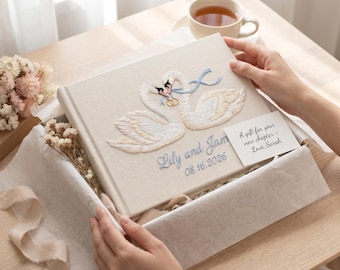 May include: A rectangular photo album with a cream-colored fabric cover featuring an embroidered design of two white swans with blue ribbons and gold rings. The text Lily and Jam 08.16.2026 is embroidered below. A small white card reads A gift for your new chapter - Love, Sarah.