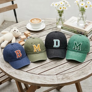 May include: Four baseball caps in navy blue, olive green, black, and green, each with a different letter. The letters are embroidered in contrasting colors: orange, yellow, light blue, and white. The caps are arranged on a wooden table with a coffee cup and flowers.