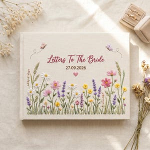 May include: A rectangular cream-colored fabric-covered book featuring embroidered wildflowers in pink, yellow, white, and purple. The text "Letters To The Bride" and "27.09.2026" are stitched in dark red, accompanied by two small embroidered butterflies and a heart.