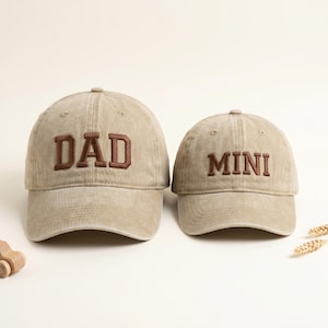May include: Two beige baseball caps, one with "DAD" embroidered in brown and the other with "MINI". The caps are made of a soft, textured fabric and have a classic, curved brim. A wooden toy car and wheat stalks are in the background.