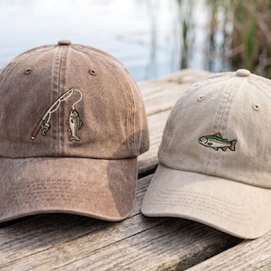 Peut inclure: Deux casquettes de baseball avec des broderies sur le thème de la pêche. L'une est marron avec une canne à pêche et un poisson, l'autre est beige avec un poisson. Les deux casquettes sont sur une surface en bois près de l'eau.