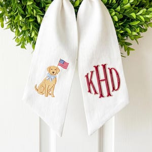 May include: Two white decorative towels. One towel features an embroidered golden retriever with a blue bow tie and an American flag. The other towel has the embroidered initials "KHD" in red.