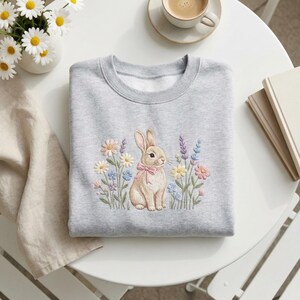 May include: A light gray sweatshirt featuring an embroidered bunny wearing a pink bow, surrounded by colorful flowers. The design includes daisies, lavender, and other wildflowers. The sweatshirt is folded neatly on a white table.