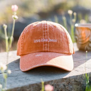 May include: An orange baseball cap with the embroidered text "love like jesus" in white. The hat is made of a soft, worn fabric and has a curved brim. The cap is sitting on a wooden surface.