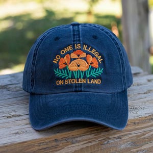 May include: A navy blue baseball cap with the embroidered text "NO ONE IS ILLEGAL ON STOLEN LAND." The cap features an embroidered design of orange flowers with green leaves.