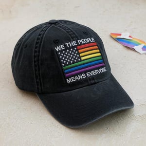 May include: Black baseball cap with a rainbow flag design and the text "WE THE PEOPLE MEANS EVERYONE." The cap features a curved brim and a fabric strap closure. The design incorporates the American flag with rainbow colors.