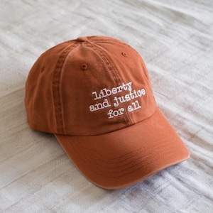 May include: A terracotta-coloured baseball cap featuring the embroidered words "Liberty and Justice for All" in white. The cap has a curved peak and a classic six-panel design, suitable for casual wear.