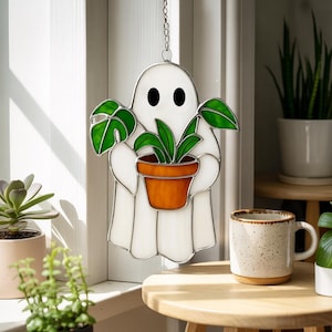 May include: A stained glass ghost holding a potted plant. The ghost is white with black eyes and a silver outline. The plant has green leaves and is in an orange pot. The piece is hanging from a chain.