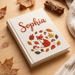 May include: A cream-colored fabric-covered journal featuring custom embroidery of the name Sophia in orange thread. The cover design includes a central fox surrounded by embroidered autumn leaves, acorns, and red-and-white spotted mushrooms.