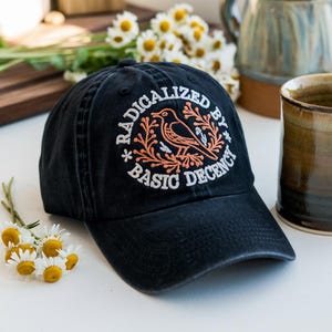 May include: A black baseball cap featuring the embroidered phrase "RADICALIZED BY BASIC DECENCY" in white. The design includes an orange bird and floral elements. The cap is on a white surface, with daisies and mugs in the background.