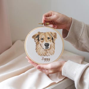 May include: Embroidered portrait of a golden retriever dog named Ziggy, displayed in a round wooden embroidery hoop. The dog's fur is detailed with shades of brown and tan, with a black nose and expressive eyes. The name "Ziggy" is stitched below.