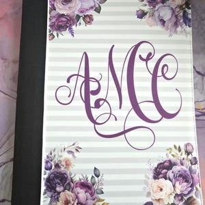 May include: A personalized notebook cover with a floral design. The cover features a gray and white striped background, adorned with purple floral arrangements in the corners. The initials "AMC" are displayed in a decorative purple font.