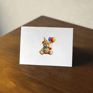 May include: A white greeting card featuring a watercolor illustration of a brown teddy bear wearing a party hat and blue scarf. The bear holds two colorful balloons. The card is on a wooden surface.