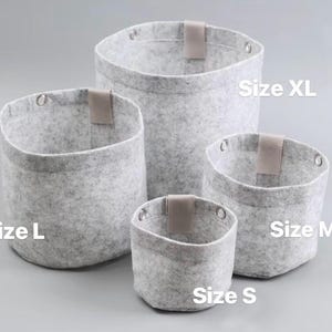 May include: Four gray felt storage baskets of varying sizes, labeled S, M, L, and XL. Each basket has a light brown leatherette tab and metal grommets. The baskets are designed for organizing and storage.