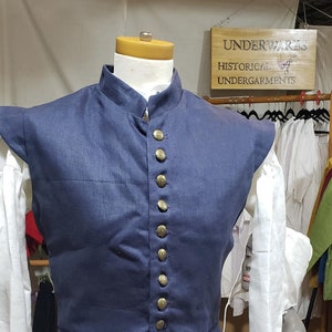 May include: A dark blue sleeveless vest with a mandarin collar and a row of brass buttons down the front. The vest has a pointed hem and is made of a linen fabric.