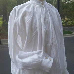 May include: A white linen shirt with a high collar and long sleeves. The shirt has a button closure at the neck and two buttons on each cuff.
