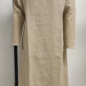 May include: A beige linen dress with long sleeves and a round neckline. The dress is floor-length and has a slit on the side. The garment is displayed on a dress form.