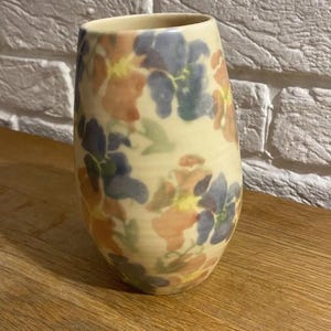 Könnte beinhalten: Eine Keramikvase mit cremefarbenem Grund, verziert mit einem Blumenmuster in Blau-, Orange- und Gelbtönen. Die Vase hat eine leicht konische Form und steht auf einer Holzoberfläche.