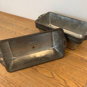 Peut inclure: Ensemble de moules à pain en métal vintage. Les moules rectangulaires ont des bords arrondis et de petites poignées. Les moules sont en métal argenté et empilés sur une surface en bois. Le texte "Regalton Bakeware England USA" est visible.