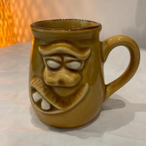 May include: A ceramic mug with a cartoonish face design. The mug is a warm, earthy brown color with white teeth and eyes. It has a curved handle and a glossy finish, perfect for coffee or tea.
