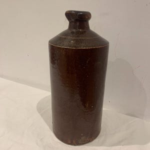 Puede incluir: Una botella alta de gres marrón con un hombro redondeado y un cuello estrecho. La botella tiene un acabado oscuro y brillante y un asa pequeña y redondeada. La botella tiene forma cilíndrica y una pequeña etiqueta rectangular en la base.