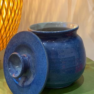 May include: A blue ceramic jar with a matching lid. The jar has a rounded body and a wide opening. The lid is slightly ajar, revealing the interior. The jar is glazed with a glossy finish, and the color is a deep, rich blue.