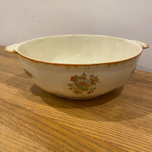 May include: Cream-coloured ceramic bowl with a floral pattern and brown trim. The bowl has two handles and a slightly distressed appearance. The floral design includes red, yellow, and green flowers. A vintage kitchenware item.
