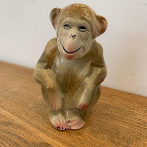 May include: A ceramic monkey figurine in a seated position. The monkey has a light brown body with a pink face, ears, and feet. It has a smiling expression and is sitting on a wooden surface.