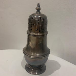 May include: A silver-colored sugar caster with a decorative pierced lid and a bulbous body. The lid is topped with a small finial. The caster sits on a round base. The overall design is ornate and vintage.