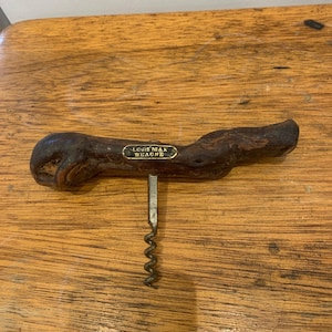 Pode incluir: Um saca-rolhas com um cabo de madeira nodosa castanho-escuro, apresentando uma pequena placa retangular de latão centrada na parte frontal. A placa tem gravada a inscrição "LOUIS MAX BEAUNE" em letras pretas. Uma espiral de metal prateado estende-se para baixo.