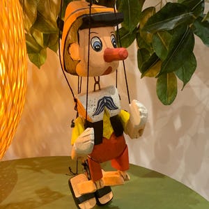 May include: A wooden Pinocchio puppet with a long red nose, wearing an orange hat, yellow shirt, and red pants. The puppet has a white mustache and is suspended by strings. The background includes green leaves and a decorative light.