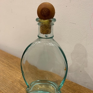 Puede incluir: Botella de vidrio transparente con cuerpo redondeado y tapón de madera. La botella tiene cierre de corcho y cuello corto. El tapón de madera tiene forma de bola redonda. La botella está sobre una superficie de madera.