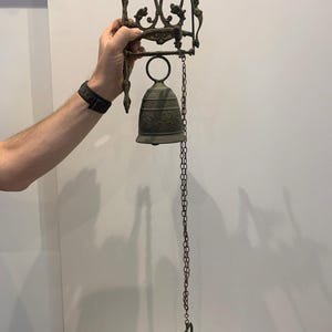 May include: A dark bronze-toned metal wall-mounted bell featuring an ornate, scrollwork bracket. The bell is suspended from the bracket by a circular ring and includes a long, thin metal chain with a decorative pull handle at the bottom end.