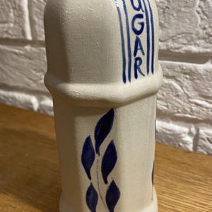 Op de afbeelding: Een witte keramische suikerpot met een blauw geschilderde "SUGAR"-opdruk en bladeren. De pot heeft een zeshoekige basis en een licht taps toelopende vorm, geschikt voor gebruik in de keuken.