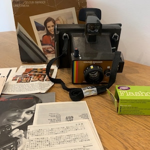 Puede incluir: Una cámara Polaroid Colour Swinger Land Camera vintage, negra con una franja arcoíris y correa negra. Acompañada de su caja original, manuales y una caja de Flashcubes Boots. La caja muestra una mujer.