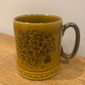 May include: Mustard-yellow ceramic mug with a silver handle. The mug features a detailed black map design with text, including the word "ON" and "PART OF SOMERSET". The base has a decorative Greek key pattern. The mug is on a wooden surface.