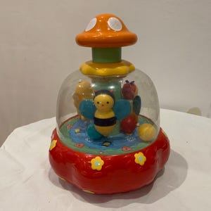 May include: A colorful children's toy with a clear dome, featuring a bee, a mushroom top, and other playful elements. The base is red with yellow flower accents, and the mushroom is orange with white spots.