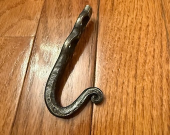 Può includere: Un gancio in metallo forgiato a mano con un ricciolo decorativo all'estremità. Il gancio è in metallo scuro e ha un aspetto rustico.