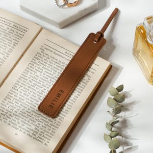 Könnte beinhalten: Ein braunes Leder Lesezeichen mit dem Namen "EMILIE" in goldenen Lettern geprägt. Das Lesezeichen liegt auf einem geöffneten Buch.