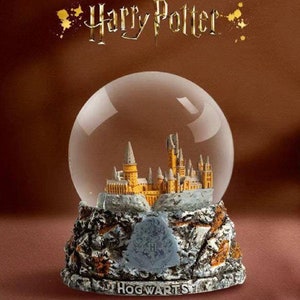 Op de afbeelding: Een glazen sneeuwbol met een gedetailleerd miniatuur van Zweinstein, omringd door een besneeuwd landschap. De basis is een rotsachtig terrein met het Zweinstein-wapen en het woord "HOGWARTS" in gouden letters gegraveerd. De sneeuwbol is een verzamelvoorwerp met het thema Harry Potter.