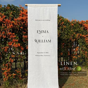 Puede incluir: Un cartel de bienvenida de boda de lino blanco con los nombres Emma y William, la fecha 8 de septiembre de 2024 y la ubicación Château Rose, Provenza. El cartel se muestra en un soporte de madera, dimensiones 56L x 69W x 61/114/170H cm.