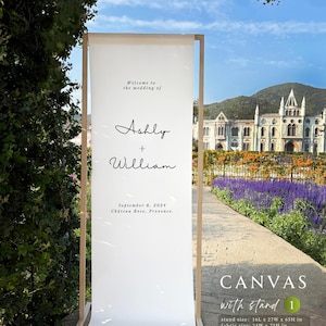 Może przedstawiać: Biały pł&oacute;cienny znak powitalny na wesele z drewnianą ramą. Na znaku widnieje napis "Welcome to the wedding of Ashly + William" i zawiera datę i miejsce. Rozmiar stojaka to 40,6 cm x 68,6 cm x 165,1 cm, a rozmiar materiału to 61 cm x 190,5 cm.